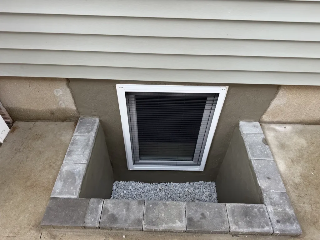 Basement window set in a block window well with paver caps and a gravel base, seen from above against gray siding