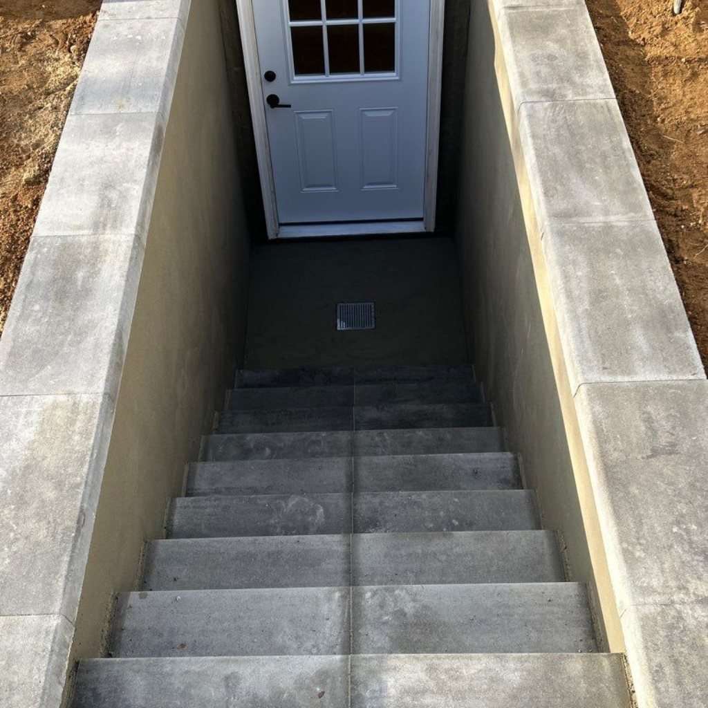 Two-wall basement walkout in Smithtown, NY with concrete steps between capped stucco walls, a floor drain on the landing and a white exterior door with a window in its upper half